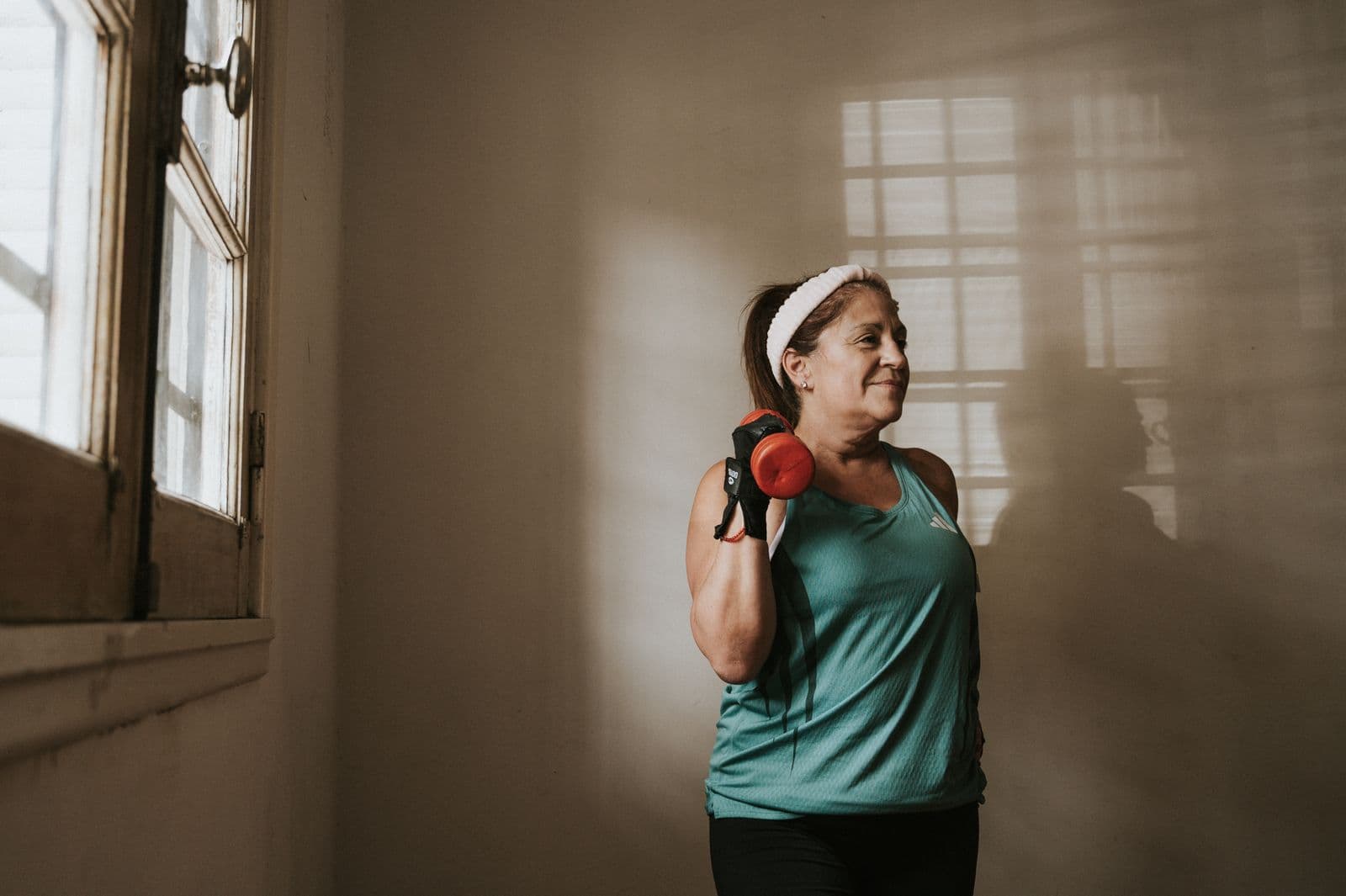 A woman in her fifties lifting a dumbbell to her shoulder in warm window light