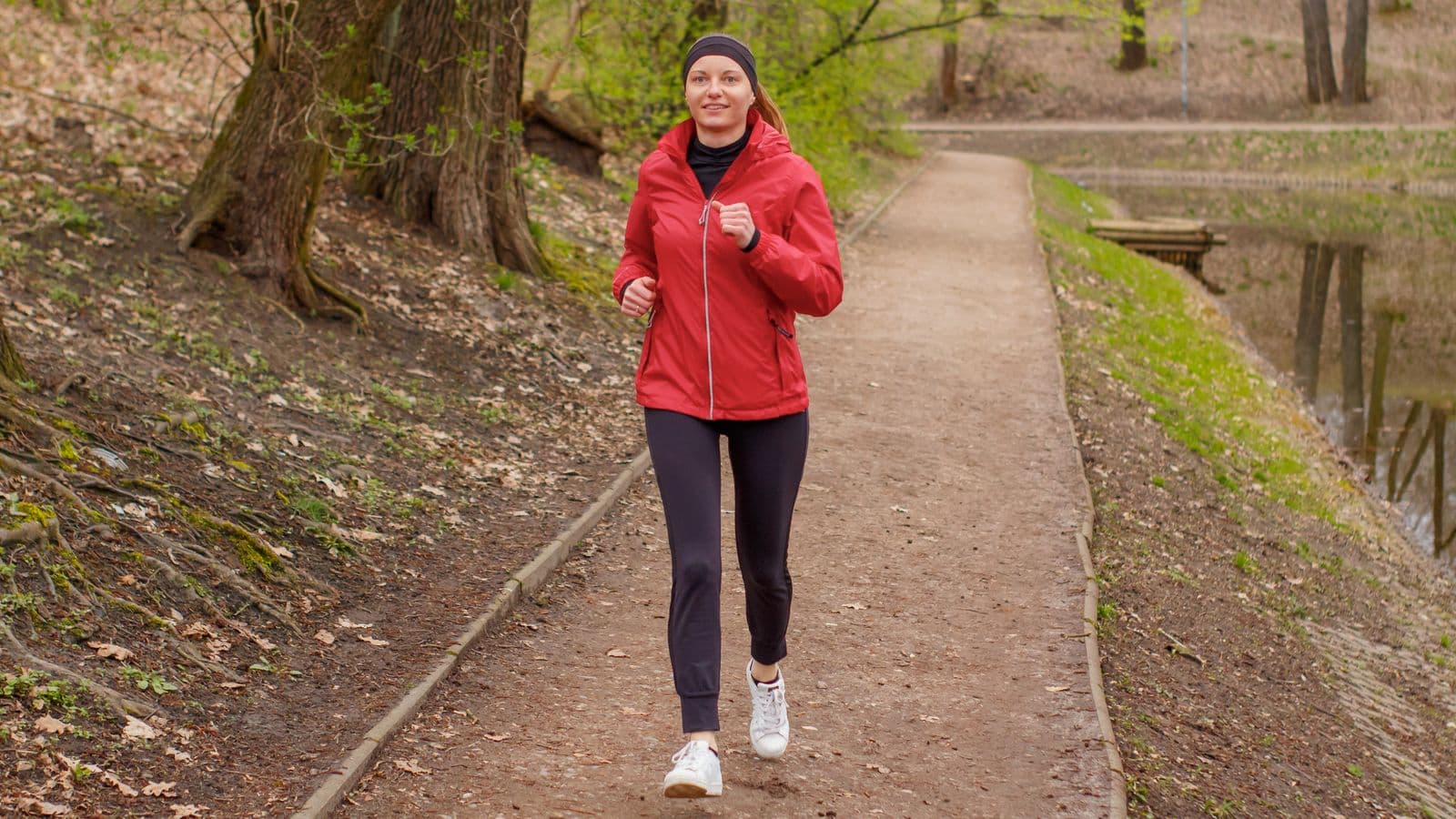 A woman in a red running jacket jogging along a dirt path through a park, beside a pond