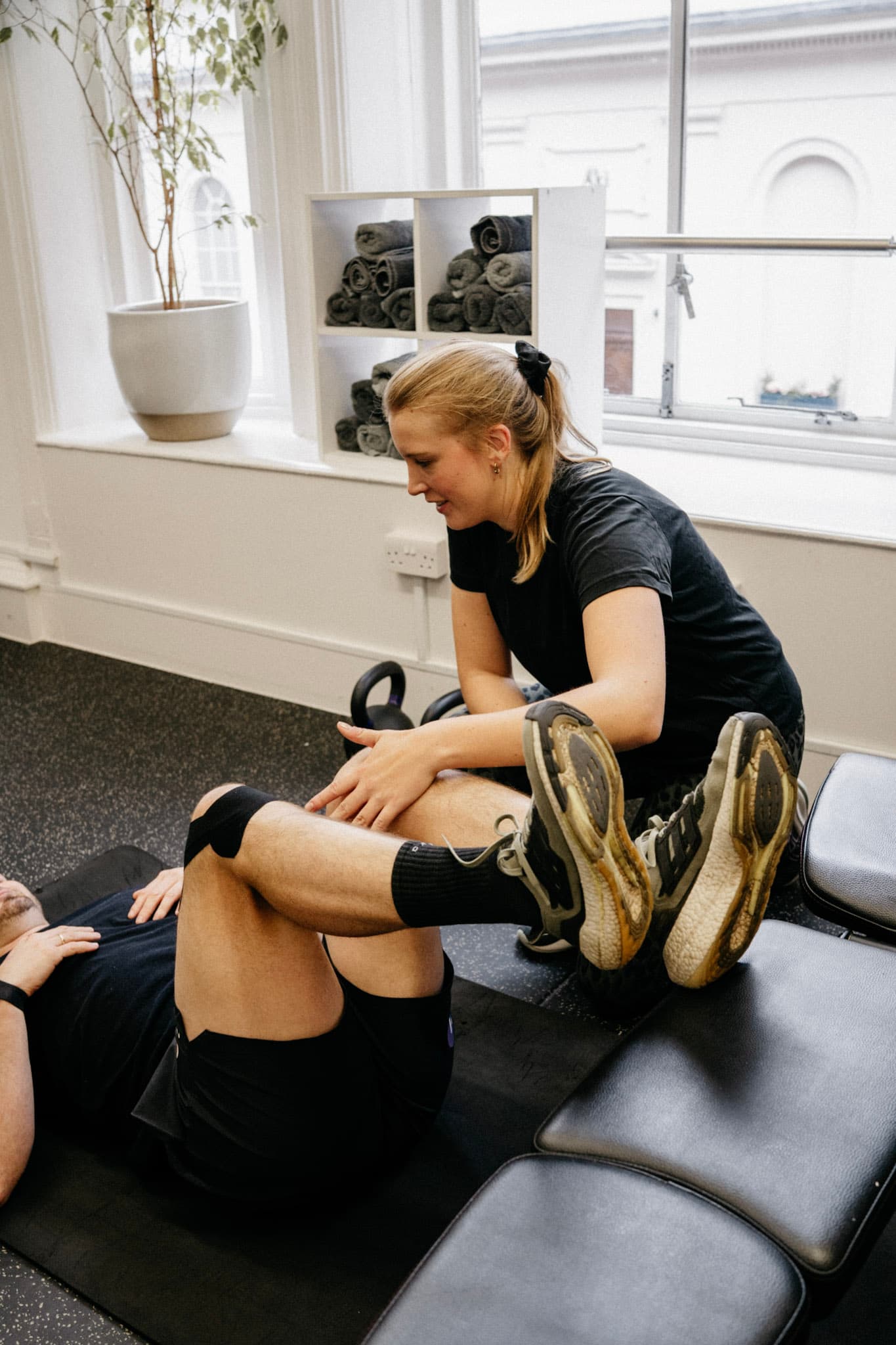 Physiotherapist assisting a client with a straight-leg raise exercise, knee taped, on the treatment bench